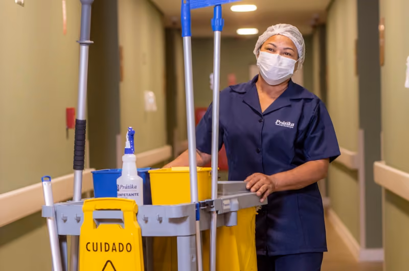 Profissional da Prátika sorrindo em corredor hospitalar, ao lado do carrinho de limpeza