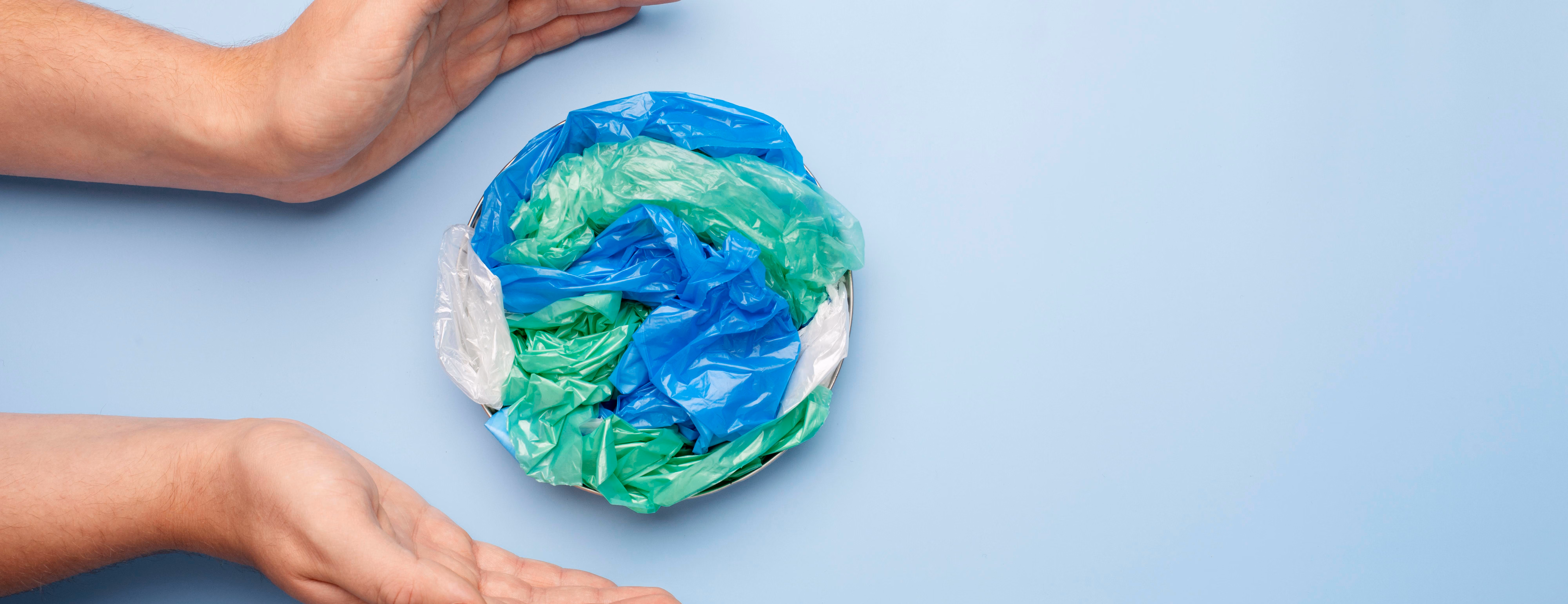 Mãos envolvem um globo feito de sacolas plásticas azuis e verdes amassadas sobre fundo azul-claro